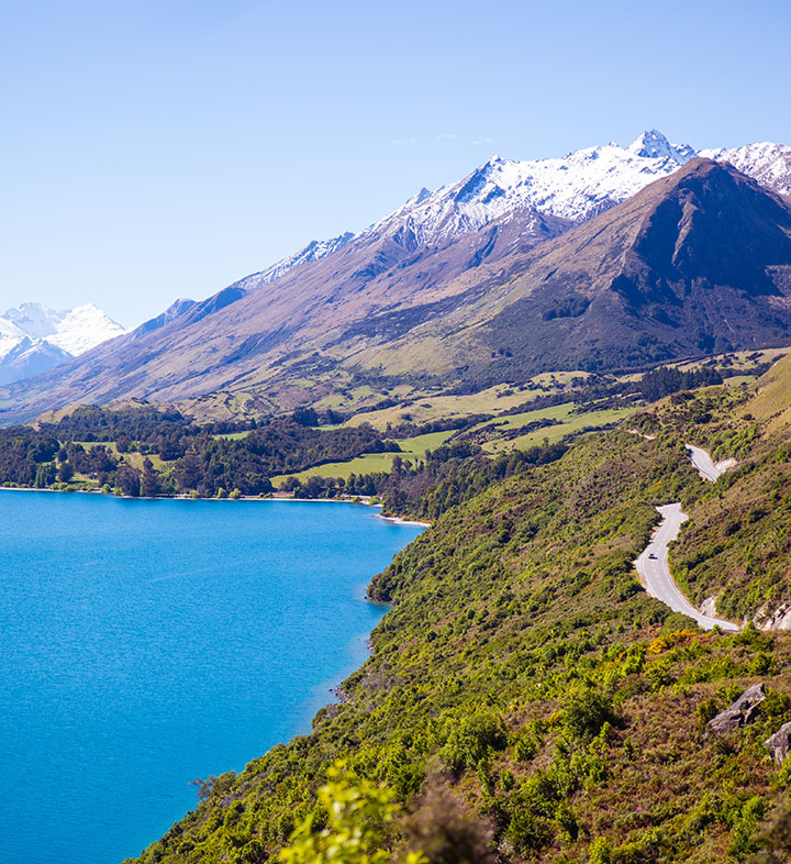 A winding road on the side of a hill above a lake in New Zealand with snowcapped mountains in the background.